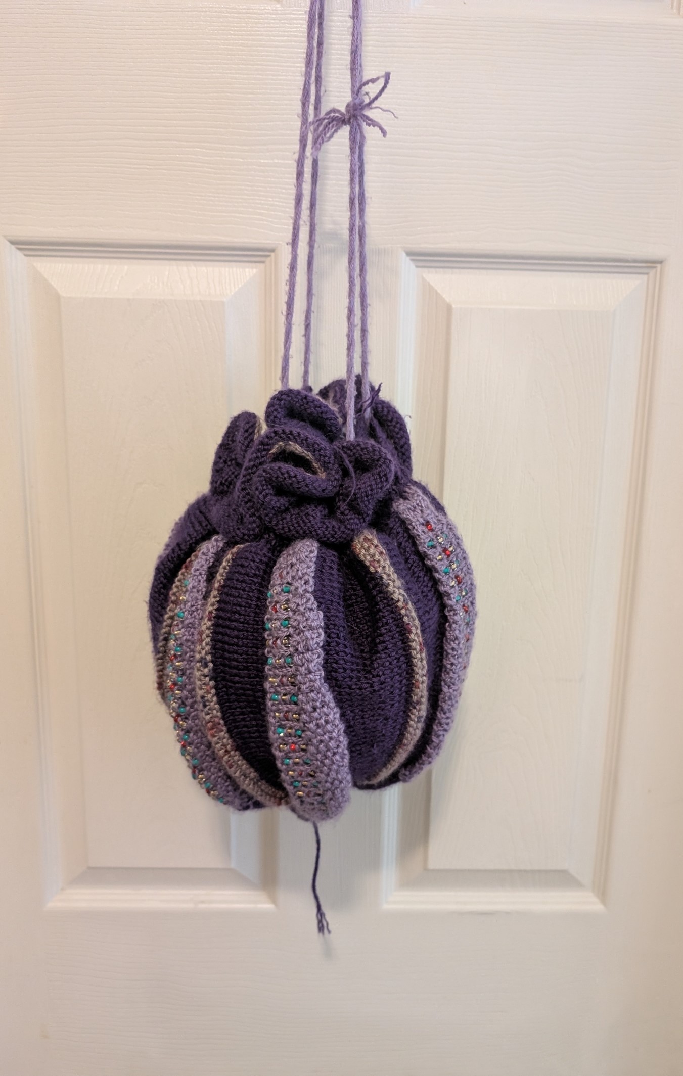  It is a circular drawstring bag made of two different shades of purple yarn and has beaded accents added to the bag. It is lined with a black acrylic fabric.