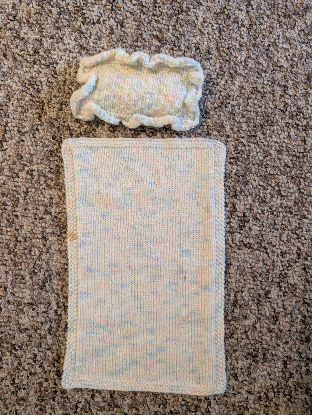 A rectangular doll pillow with a ruffled edging made of blue, yellow, pink and white yarn. There is also a doll blanket made out of the same yarn. It is rectangular as well, but does not feature a ruffled edging; instead, it has folded edging. 