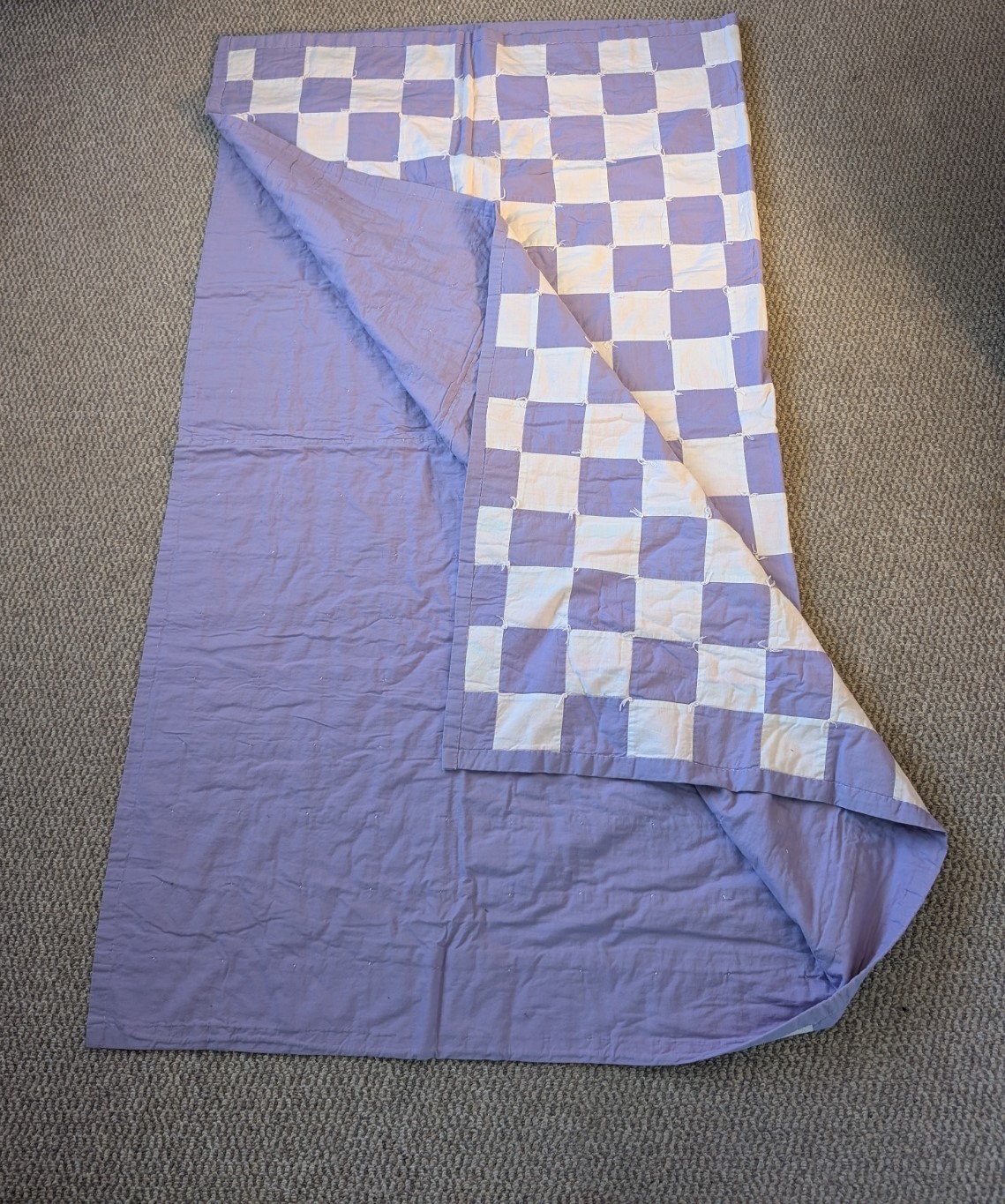 A purple and white quilt. It features patches of white and purple fabric on the front and a purple back. The two pieces of the quilt are held together by the hand-tied quilting method.