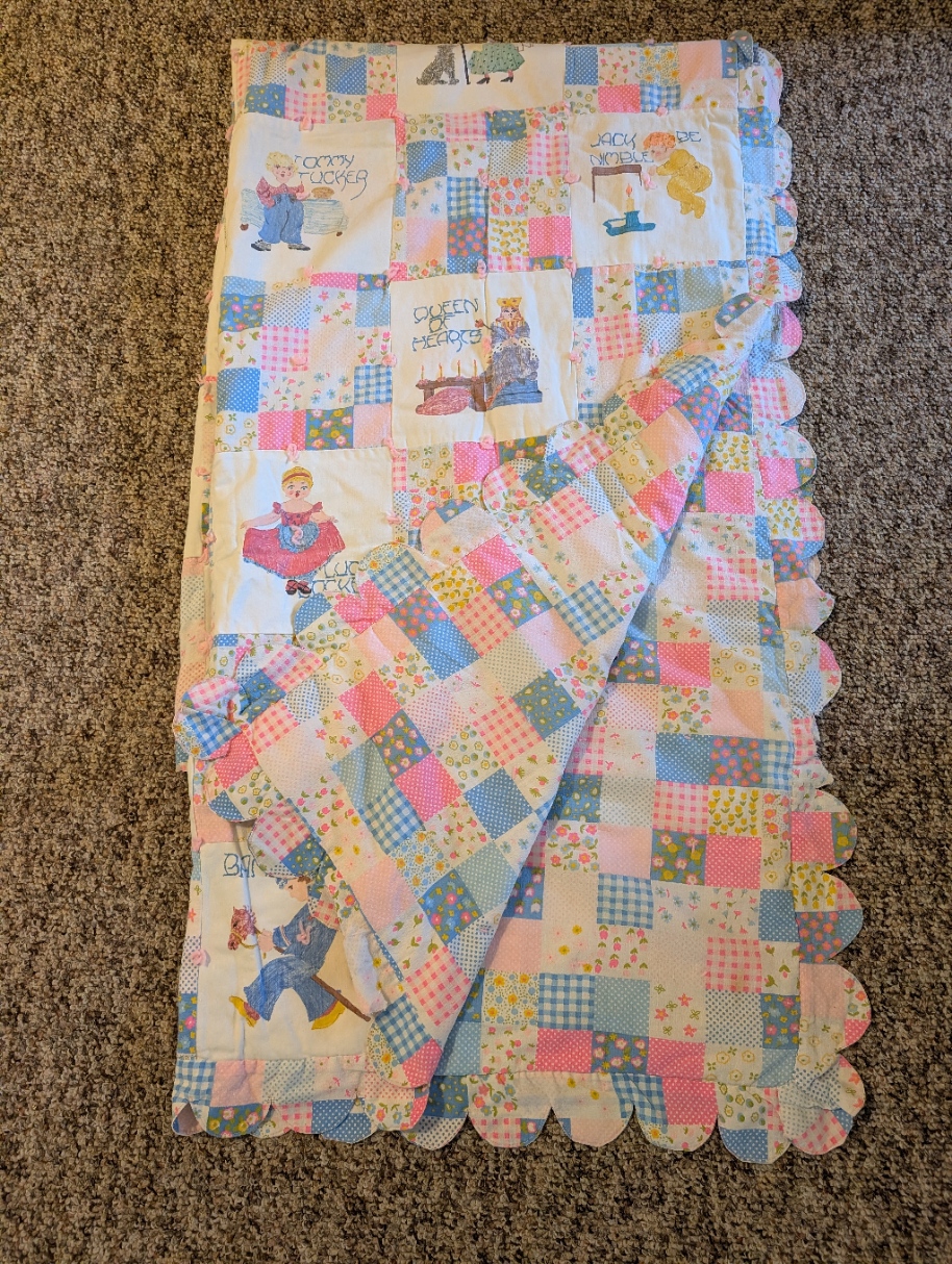 A quilted blanket featuring a patchwork of flower fabric and a white fabric with nursery rhyme characters drawn on using fabric paint. The quilt is finished with a crescent fabric edging around the entire blanket, except for the top, where a blanket stitch is used. 