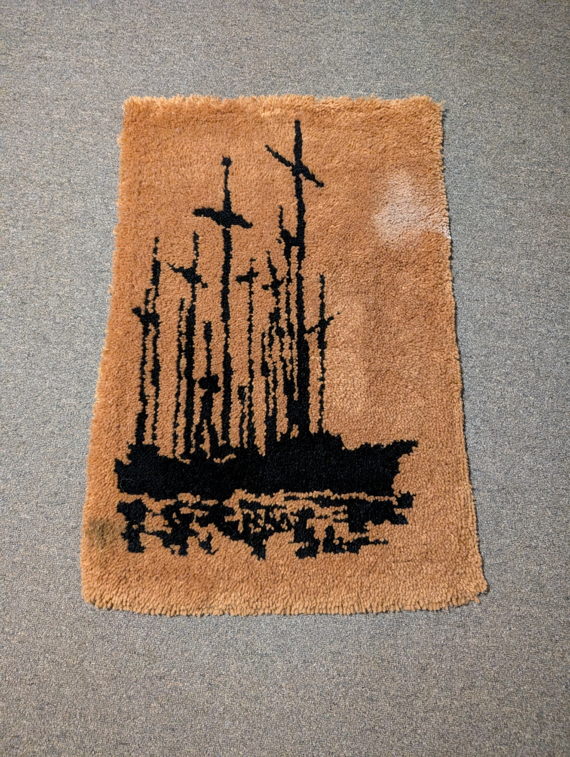 A rectangular rug that is meant to hang off the wall. The background is brown, and in the center of the piece is a ship made with dark blue yarn. It has a few stains on the sides of the rug.