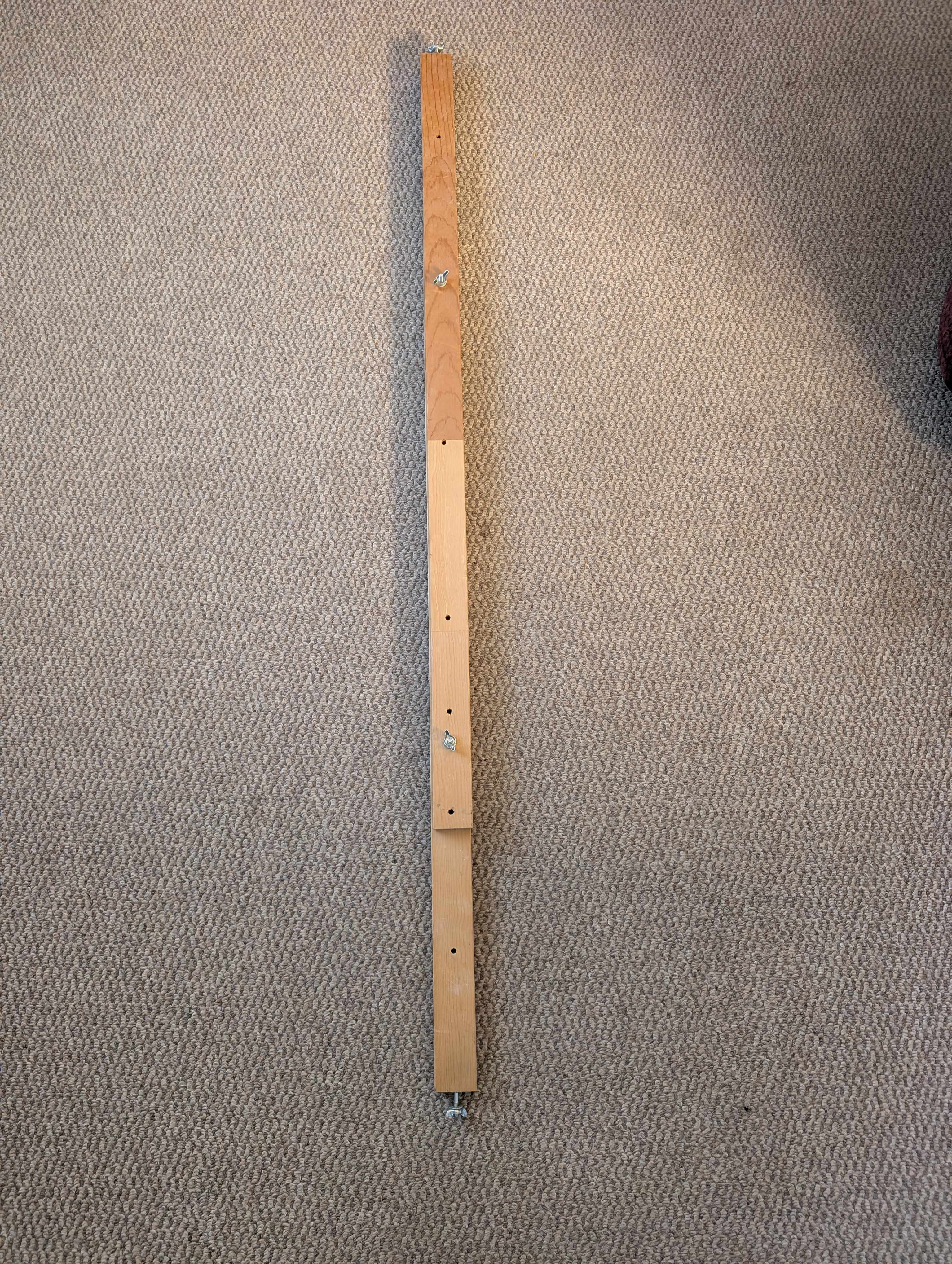 A long piece of wood lies flat on a brown carpet, lengthwise to the camera. It consists of three pieces of wood stacked on top of each other and held together by butterfly thumb screws. These screws also hold the piece together at the ends of the wood stacks.