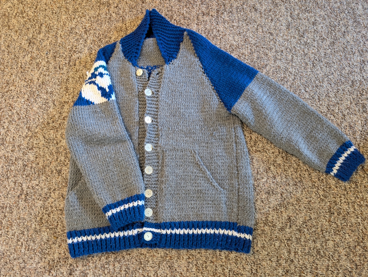 Blue Jays Cardigian  A grey, blue, and white knitted cardigan with a hand-designed tapestry knitting pattern on the back. There is a Blue Jays logo on the right sleeve and my brother's last name, which a piece of folded paper has covered, and the number 76, his baseball number.
