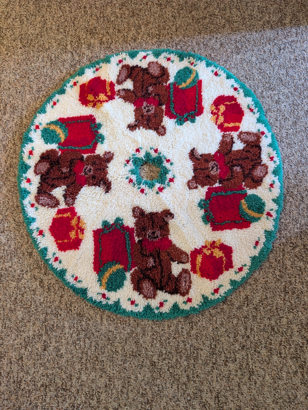 Christmas Rug Hooking A circular tree skirt with a white base and green edging, and small holly berries going around the edging. It features a picture of a bear wearing a bow, accompanied by two red Christmas presents and a ball on either side. This same image repeats around the skirt four times.